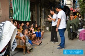 Two people talking to many street children, with a tag reading "Anna Natividad | ITE ET DOCETE" at the corner