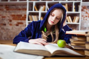 A hooded person reading some books in a library with an apple on top of a book