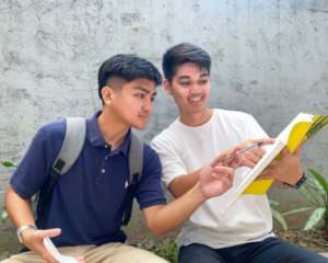 Two students outside, with one teaching the other using a book